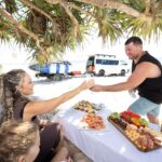 A man and woman exchange drinks at a picnic table set with seafood, fruit, and snacks on a sandy beach under leafy trees, with vans and surfboards in the background.