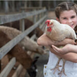 A girl smiles while holding a white chicken in her arms at an outdoor animal enclosure, with other animals visible in the background.