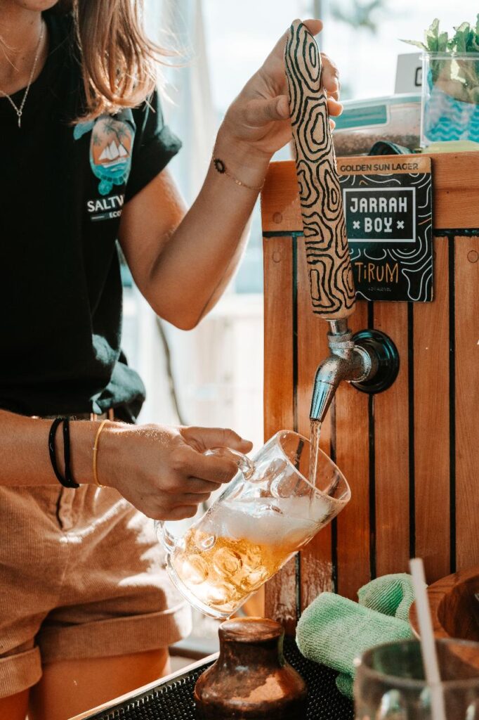 A person pours beer from a tap labeled "Jarrah Boy" into a glass mug at an outdoor setting.