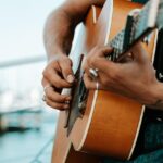 A person wearing rings and sleeveless clothing is playing an acoustic guitar outdoors near the water.