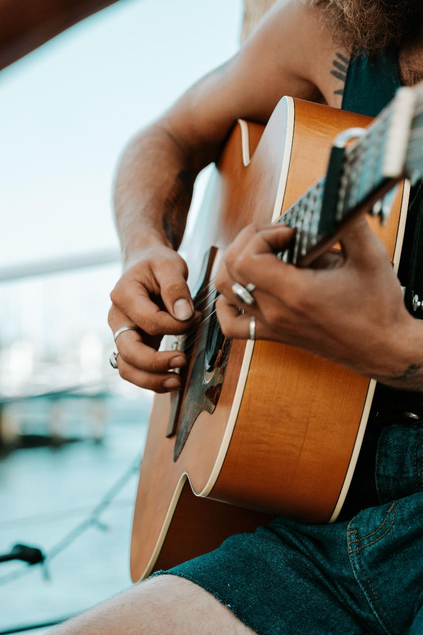 A person wearing rings and sleeveless clothing is playing an acoustic guitar outdoors near the water.