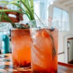 Two tall glasses of iced orange-colored fruit drink with berries and green garnish sit on a wooden table, with a blurred outdoor background.
