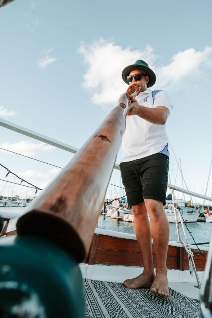 A person standing barefoot on a boat deck plays a didgeridoo, with boats and a marina visible in the background.