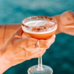 A person holding a cocktail in a coupe glass with a chocolate-rimmed edge, against a blurred blue background.