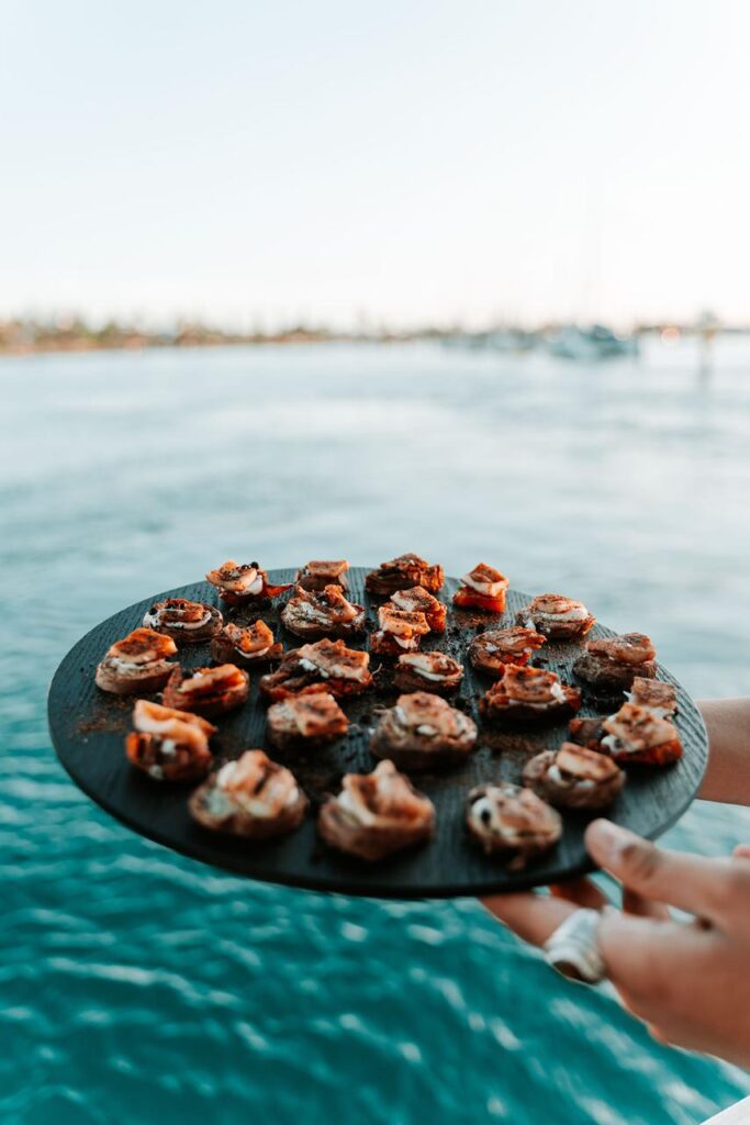 A hand holds a round tray with bite-sized appetizers above blue water, with a blurred shoreline and boats in the background.