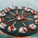 A black round platter with crackers topped with white cream and a reddish garnish, arranged in a circle, with thin strips of food radiating from the center.