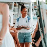 A woman in a white shirt and denim shorts holds a tray of snacks while smiling at a group of people on a boat.