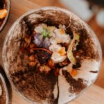 A bowl made from a coconut shell containing granola, dried fruit, and edible flowers, placed on a wooden surface.