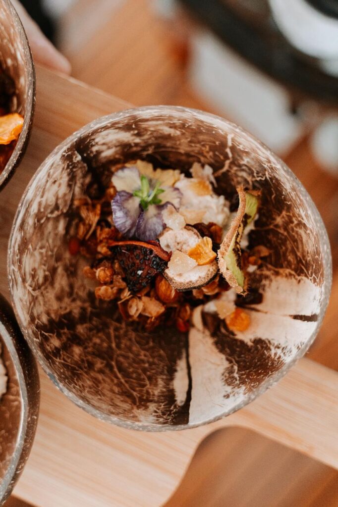 A bowl made from a coconut shell containing granola, dried fruit, and edible flowers, placed on a wooden surface.