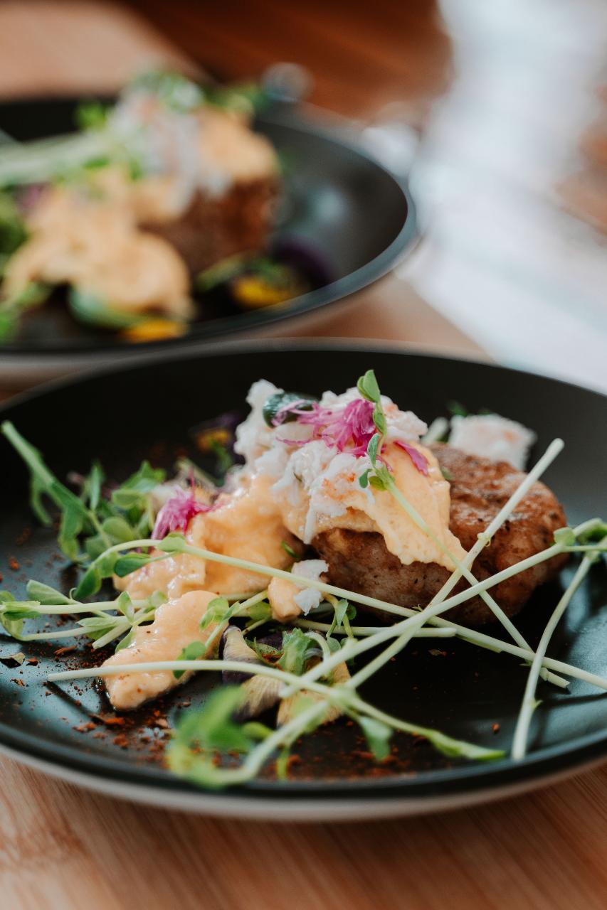 A close-up of a plated dish featuring a patty topped with a creamy yellow sauce, microgreens, and edible flowers, served on a black plate.
