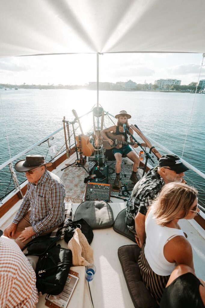 A group of people sit on a boat as a musician plays guitar and sings; the boat is on calm water with buildings visible in the background.