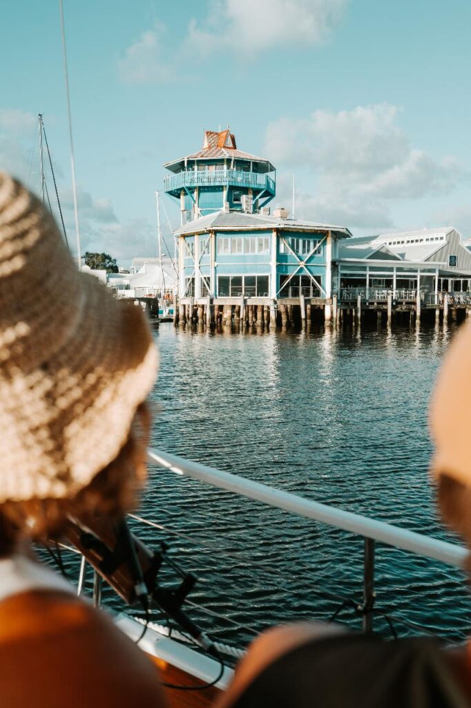 Two people on a boat look toward a blue waterfront building with large windows and a tower, under a partly cloudy sky.