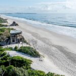 Aerial view of a sandy beach with waves, coastal vegetation, and an old concrete structure near the shoreline.