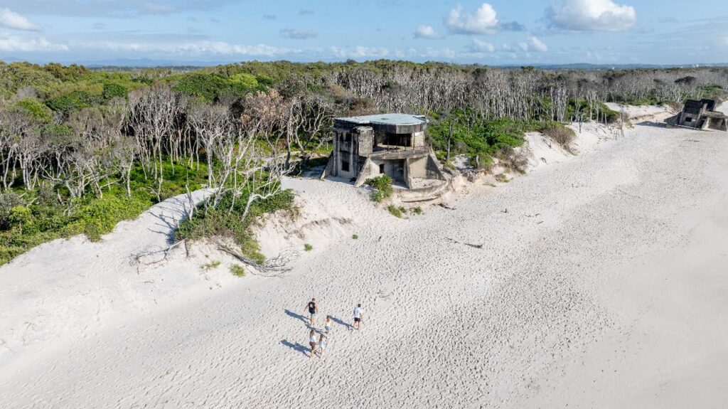 A group of people walks on a sandy beach near old concrete bunkers and sparse coastal trees under a partly cloudy sky.