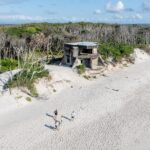 A group of people walks on a sandy beach near old concrete bunkers and sparse coastal trees under a partly cloudy sky.