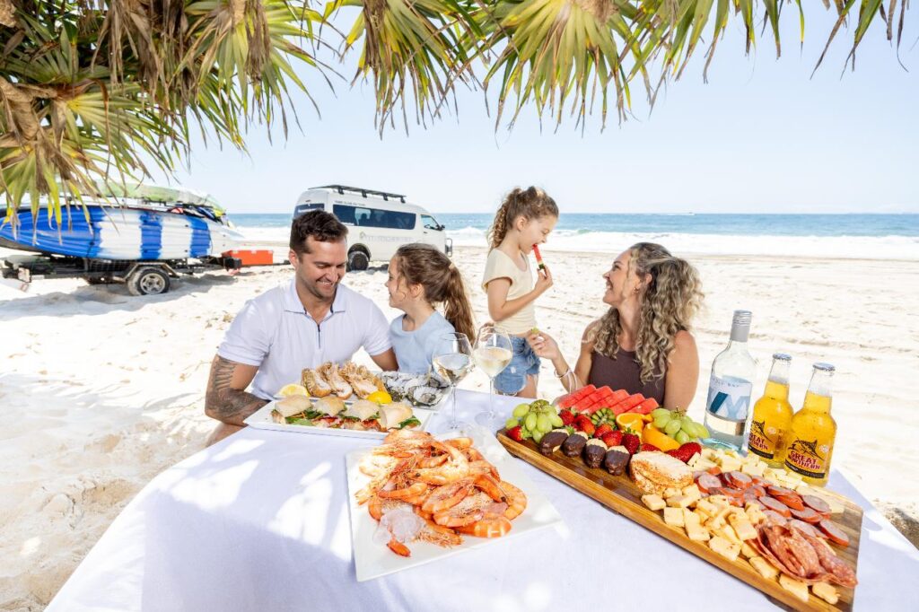 A family sits at a table on a sandy beach, enjoying a seafood and fruit picnic under palm trees, with a van and surfboards in the background near the ocean.
