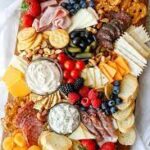 A charcuterie board with assorted cheeses, meats, crackers, bread, fresh berries, tomatoes, nuts, dried apricots, and two bowls of dip.