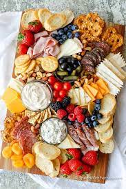 A charcuterie board with assorted cheeses, meats, crackers, bread, fresh berries, tomatoes, nuts, dried apricots, and two bowls of dip.