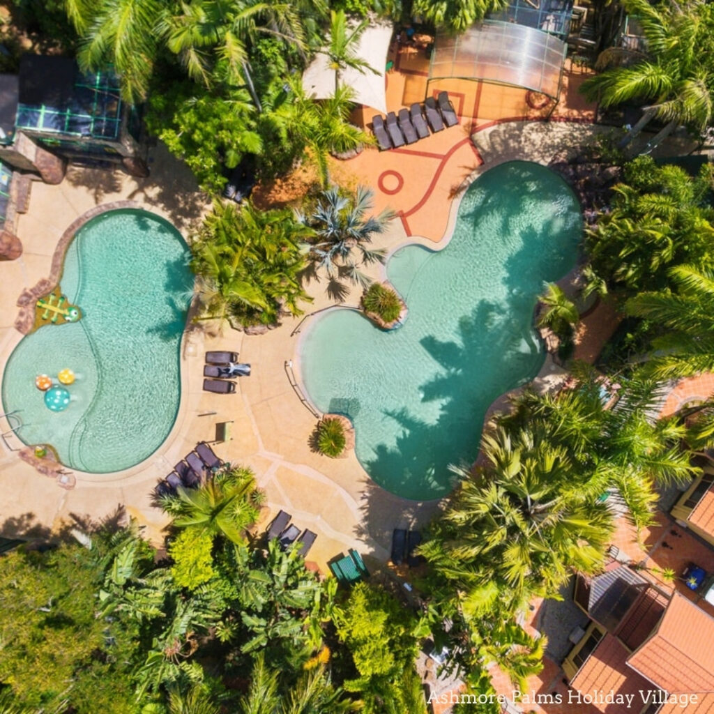 Aerial view of two outdoor swimming pools surrounded by palm trees, lounge chairs, and lush greenery at a resort.