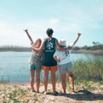 Three people stand with their backs to the camera at the edge of a lake, arms raised and making hand signs, under a blue sky with greenery and water in the background.