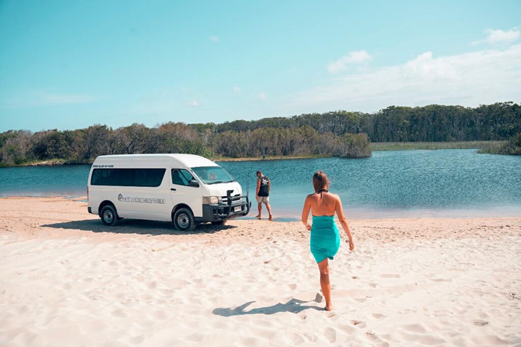 A woman in a blue dress walks toward a white van parked on a sandy beach by a body of water, with trees visible in the background.