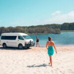 A woman in a blue dress walks toward a white van parked on a sandy beach by a body of water, with trees visible in the background.