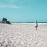 A person walks alone on a sandy beach near the shoreline, with ruins of a concrete structure visible in the distance under a clear blue sky.