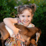 A young girl with pigtails, wearing a floral dress, smiles while holding a brown and white baby goat on her lap outdoors.