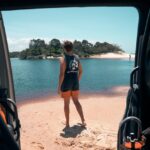 Person in a sleeveless shirt and shorts stands on a sandy beach by a body of water, viewed from inside a vehicle with equipment visible in the foreground.