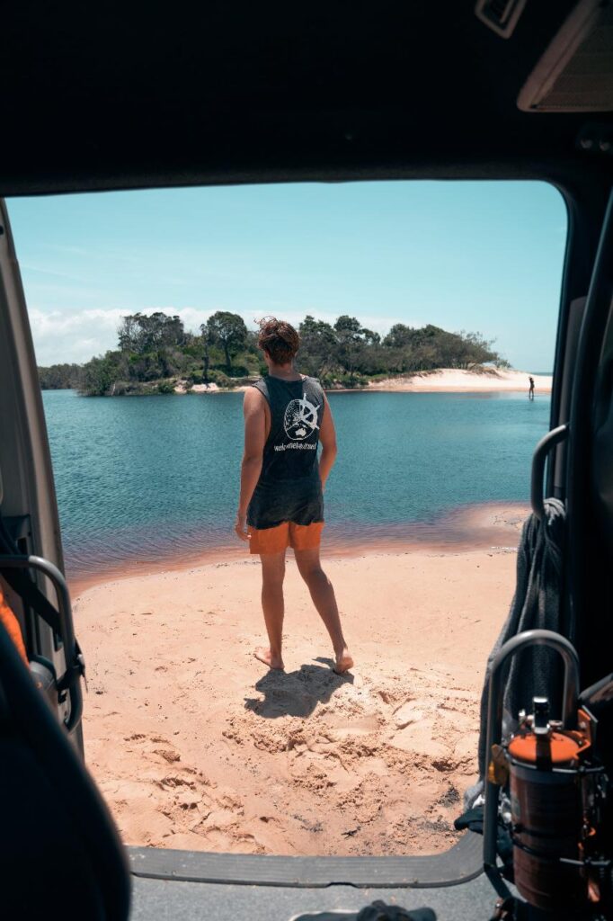 Person in a sleeveless shirt and shorts stands on a sandy beach by a body of water, viewed from inside a vehicle with equipment visible in the foreground.
