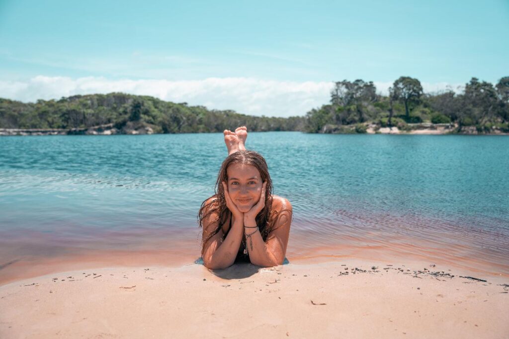 A girl lies on her stomach at the edge of a lake, resting her chin on her hands, with trees and blue sky in the background.