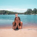 A girl lies on her stomach at the edge of a lake, resting her chin on her hands, with trees and blue sky in the background.