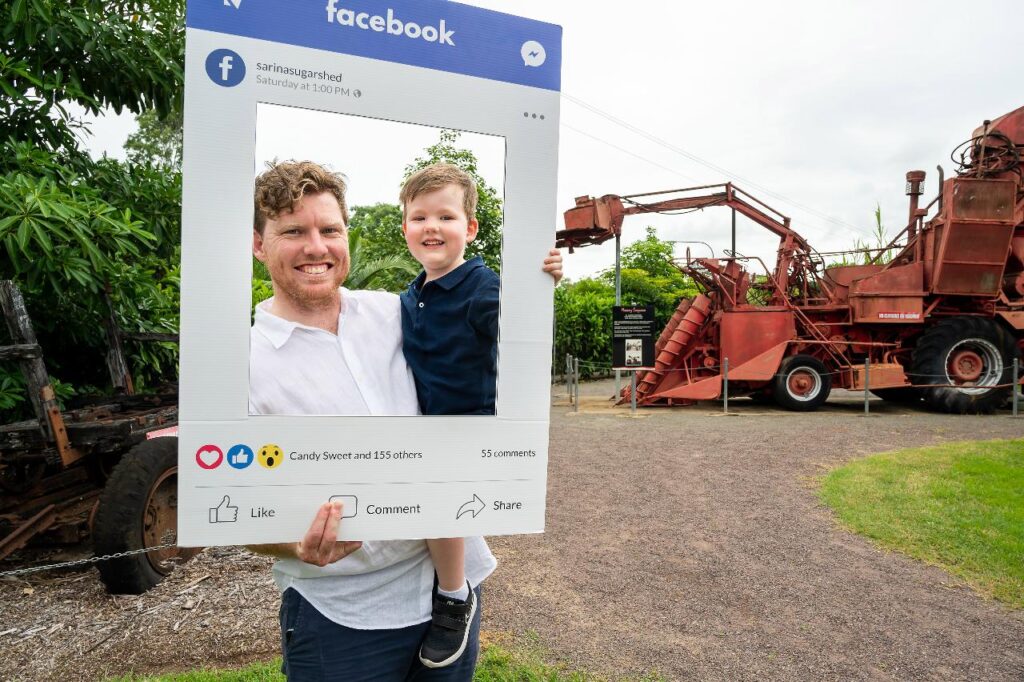 A man holds a young boy while both pose behind a large mock Facebook photo frame; old farming machinery is visible in the background outdoors.
