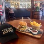A burger and fries served on a wooden tray with dipping sauce, next to a black cap on a table in a bar-style restaurant.
