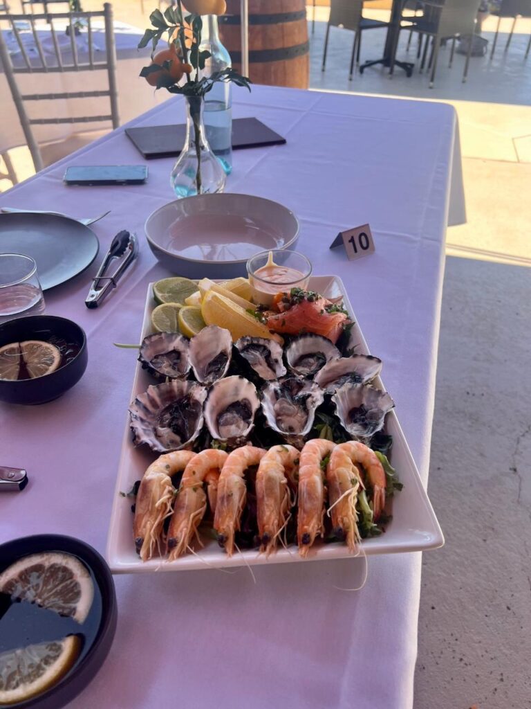 A rectangular plate with oysters, shrimp, lemon wedges, and salmon sits on a white tablecloth, surrounded by dishes, cutlery, and a vase with flowers.