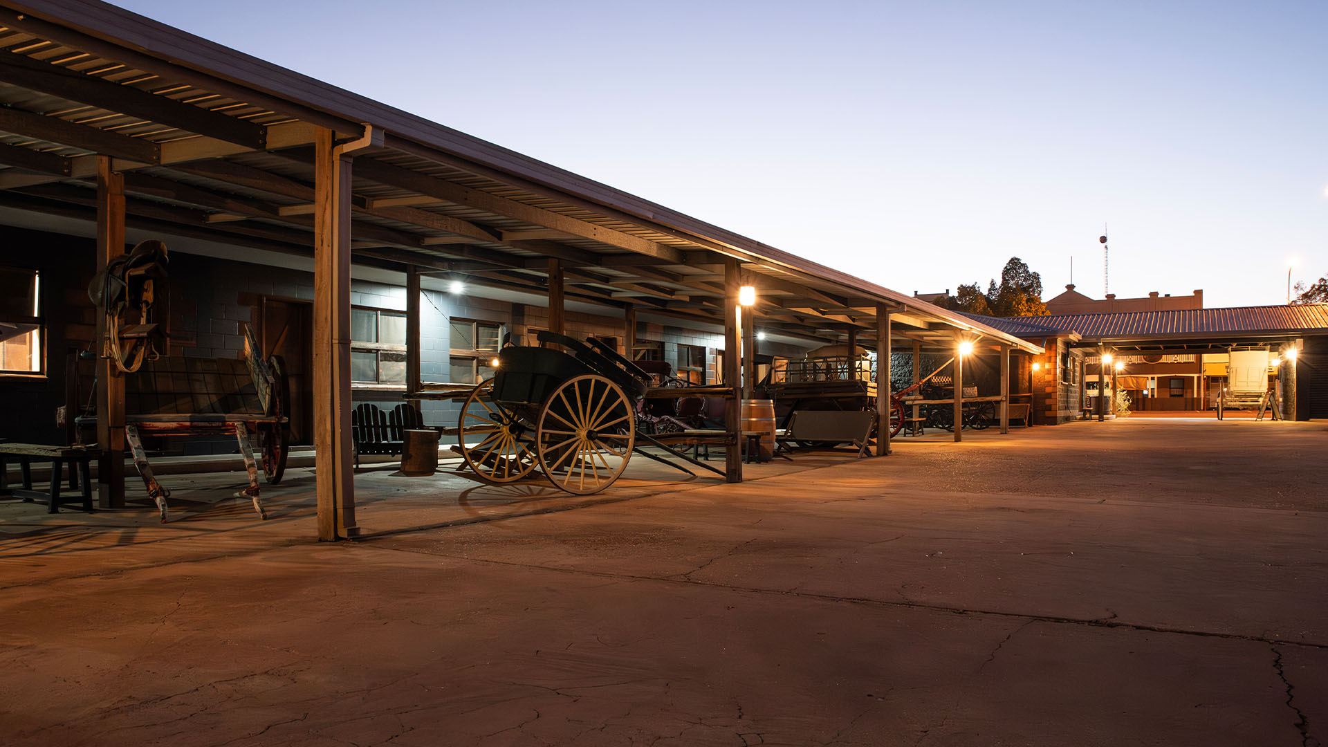A covered outdoor area at dusk displays old-fashioned carriages and equipment, illuminated by several lights mounted on the structure.