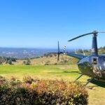 A black helicopter is parked on a grassy field with rolling hills and a clear blue sky in the background.