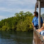 A group of people wearing hats stand on a boat, looking out at a river surrounded by green trees under a blue sky.