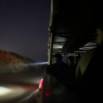 People ride in an open vehicle at night, looking out into the dark as headlights and a beam of light illuminate the road ahead.