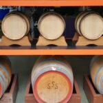Several wooden barrels are stored on metal shelves, with six smaller barrels on the top row and three larger barrels marked “SSS” on the bottom row.
