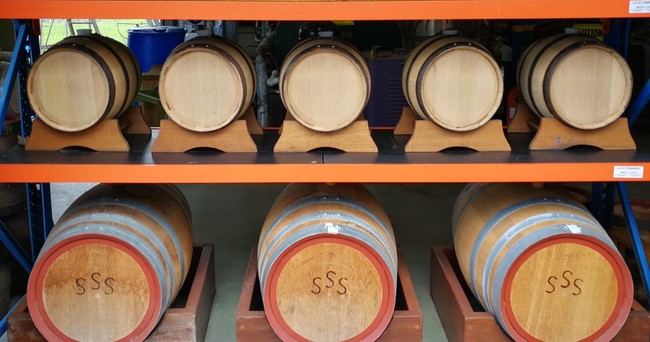 Several wooden barrels are stored on metal shelves, with six smaller barrels on the top row and three larger barrels marked “SSS” on the bottom row.