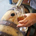A person fills a glass with amber liquid from a metal spout attached to a wooden barrel.