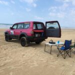 A pink SUV with its rear door open is parked on a sandy beach next to a folding table, two chairs, and camping items. The ocean and sky are in the background.