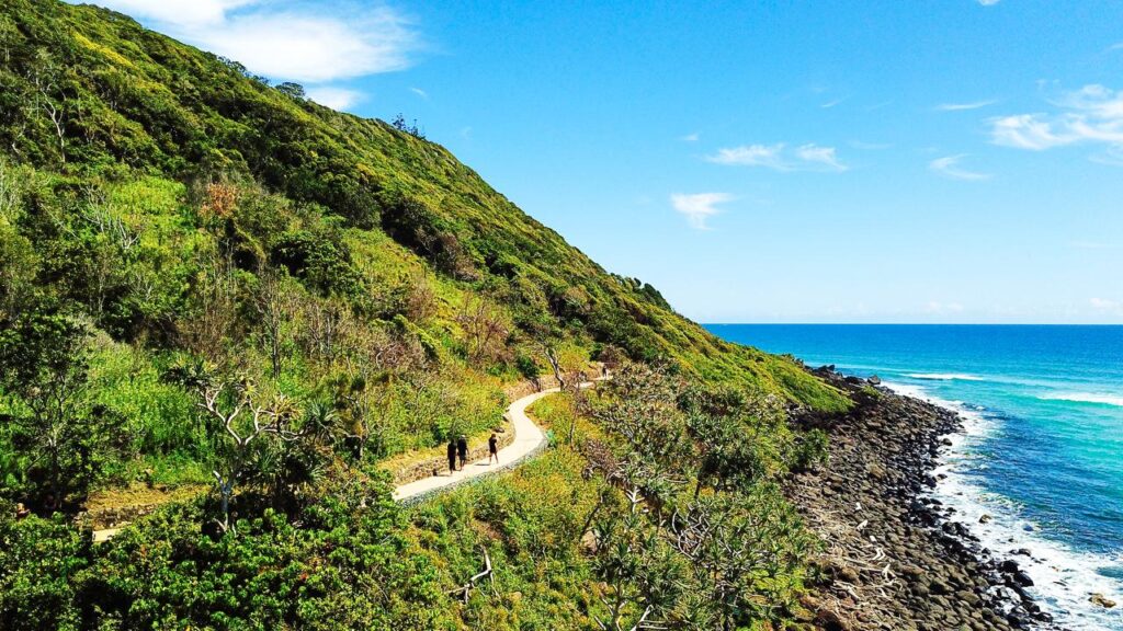 A winding coastal path curves along a green hillside beside the blue ocean, with a few people walking under a clear, sunny sky.