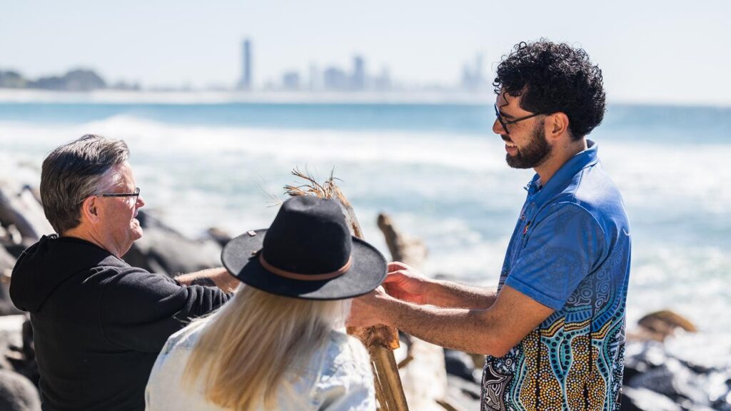 Three people stand by the ocean, holding wooden sticks and talking. The city skyline is visible in the background.