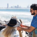 Three people stand by the ocean, holding wooden sticks and talking. The city skyline is visible in the background.
