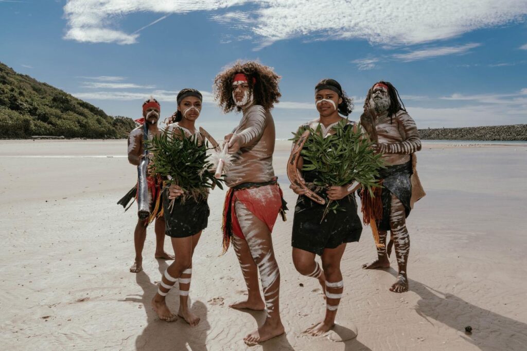 Five people in traditional attire with body paint stand on a sandy beach holding green plants, with blue sky and greenery in the background.