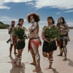 Five people in traditional attire with body paint stand on a sandy beach holding green plants, with blue sky and greenery in the background.