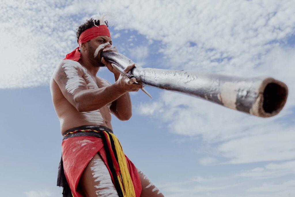 A person with body paint and traditional attire plays a didgeridoo outdoors under a partly cloudy sky.