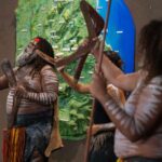 Three people with traditional body paint and attire hold boomerangs, performing in front of a wall decorated with green and blue elements.